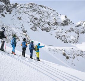 Eine Gruppe von Skifahrern steht auf einem schneebedeckten Berg. Im Hintergrund sind majestätische Berggipfel zu sehen.