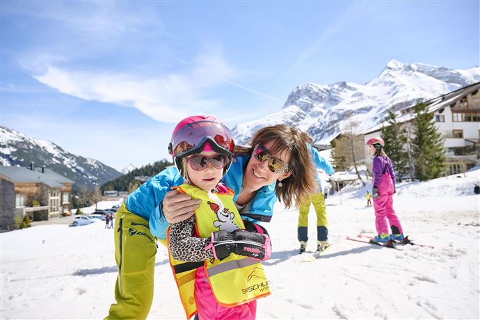 Eine fröhliche Mutter umarmt ihr Kind auf einer Skipiste. Im Hintergrund sind schneebedeckte Berge und andere Skifahrer zu sehen.