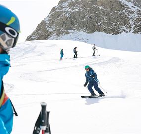 Ein Skifahrer fährt auf einer verschneiten Piste. Im Hintergrund sind weitere Personen beim Skifahren zu sehen.