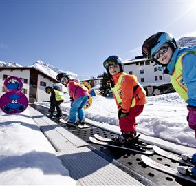 Eine Gruppe von Kindern in bunten Skianzügen steht auf Skiern im Schnee. Im Hintergrund sind schneebedeckte Berge und ein Haus zu sehen.