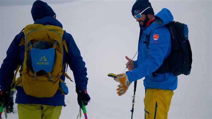Zwei Bergsteiger stehen im Schnee und betrachten ein Gerät. Sie tragen winterliche Outdoor-Bekleidung und haben Wanderausrüstung dabei.