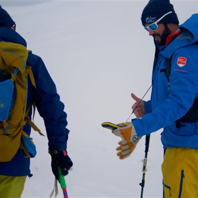 Zwei Bergsteiger stehen im Schnee und betrachten ein Gerät. Sie tragen winterliche Outdoor-Bekleidung und haben Wanderausrüstung dabei.