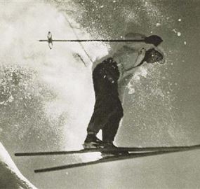 Ein Skifahrer springt über eine schneebedeckte Piste. Der Schnee spritzt beim Sprung in die Höhe.