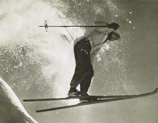Ein Skifahrer springt über eine schneebedeckte Piste. Der Schnee spritzt beim Sprung in die Höhe.