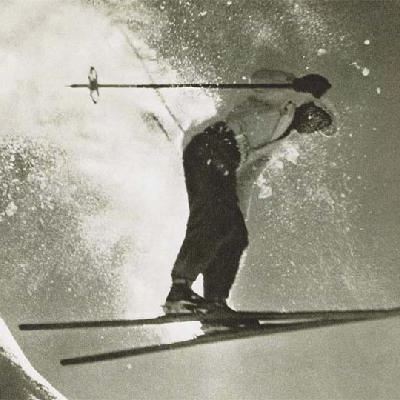 Ein Skifahrer springt über eine schneebedeckte Piste. Der Schnee spritzt beim Sprung in die Höhe.