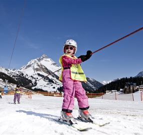 Ein kleines Kind in pinker Skiausrüstung fährt auf einem Skihang. Im Hintergrund sind schneebedeckte Berge und ein blauer Himmel zu sehen.