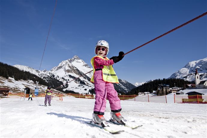 Ein kleines Kind in pinker Skiausrüstung fährt auf einem Skihang. Im Hintergrund sind schneebedeckte Berge und ein blauer Himmel zu sehen.