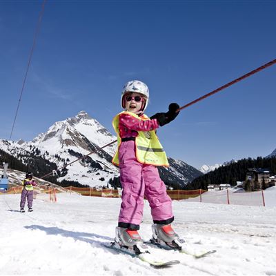 Ein kleines Kind in pinker Skiausrüstung fährt auf einem Skihang. Im Hintergrund sind schneebedeckte Berge und ein blauer Himmel zu sehen.