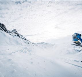 Ein Skifahrer springt durch den Schnee in den Bergen. Der Himmel ist bewölkt und die Landschaft ist winterlich und malerisch.