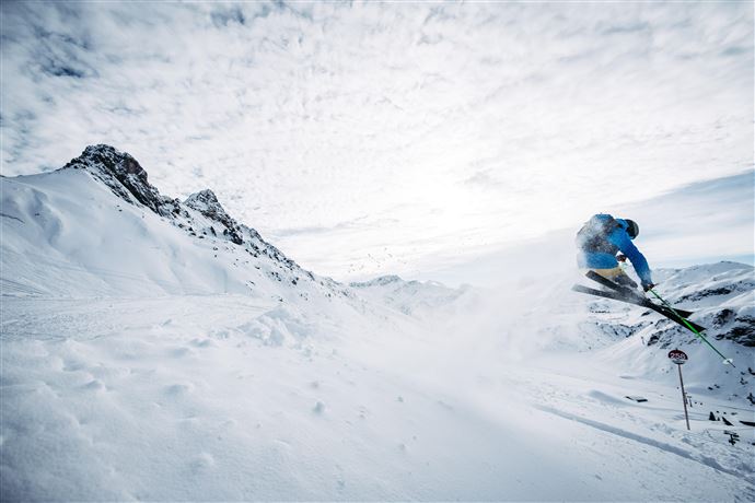 Ein Skifahrer springt durch den Schnee in den Bergen. Der Himmel ist bewölkt und die Landschaft ist winterlich und malerisch.