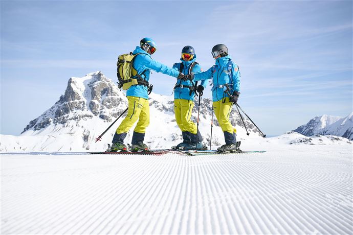 Drei Skifahrer stehen auf einer schneebedeckten Piste und halten sich gegenseitig bei den Händen. Im Hintergrund sind majestätische Berge und ein klarer Himmel zu sehen.