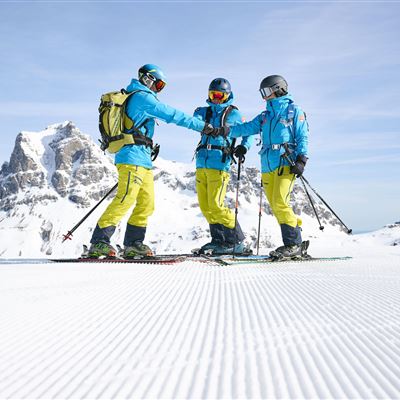 Drei Skifahrer stehen auf einer schneebedeckten Piste und halten sich gegenseitig bei den Händen. Im Hintergrund sind majestätische Berge und ein klarer Himmel zu sehen.