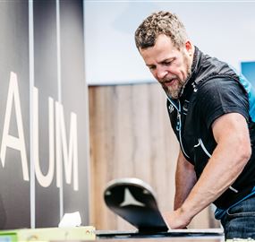 Ein Mann arbeitet an einem Tisch, während er mit einem Sportgerät beschäftigt ist. Im Hintergrund steht das Wort "RAUM" an einer Wand.