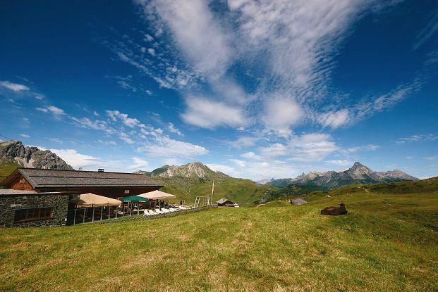Eine malerische Almhütte umgeben von grünen Wiesen und hohen Bergen. Der Himmel ist blau mit einigen Wolken.