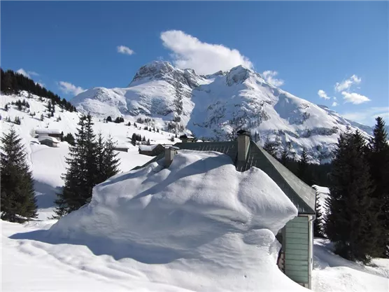 A snow-covered mountain landscape with tall mountains and a clear sky. In the foreground, there is a roof hidden beneath the snow.