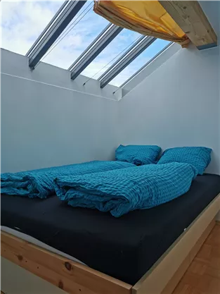 Ein gemütliches Schlafzimmer mit einem Doppelbett. Die Bettwäsche ist in hellblau, und große Fenster lassen viel Licht herein.