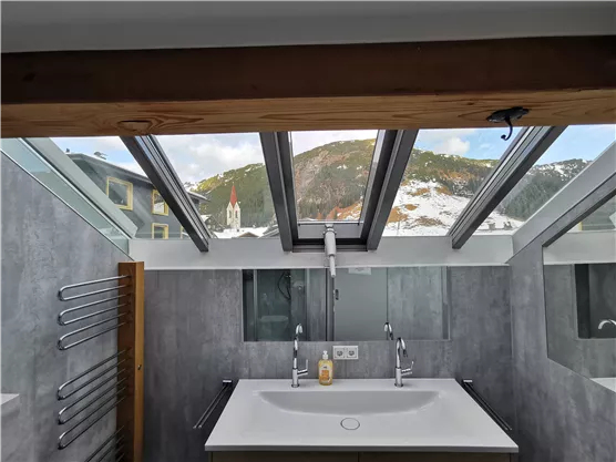 Ein modernes Badezimmer mit einem großen Waschbecken und einem Fensterdach. Im Hintergrund sind schneebedeckte Berge und eine Kirche sichtbar.