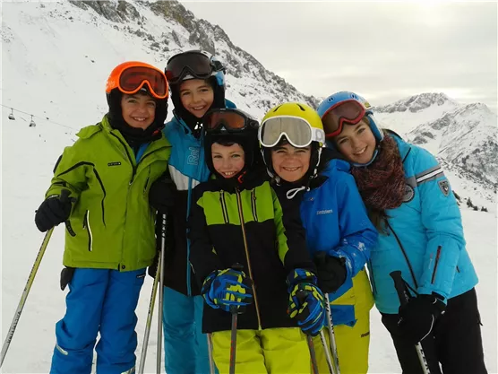 A group of five children stands on a snowy slope wearing ski gear. In the background, there are mountains and a cloudy sky.
