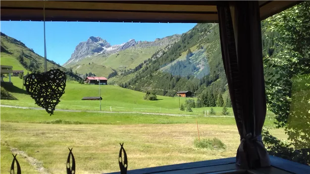 Eine malerische Alpenlandschaft mit grünen Wiesen und Bergen im Hintergrund. Einige Hütten sind zwischen den Bäumen sichtbar.