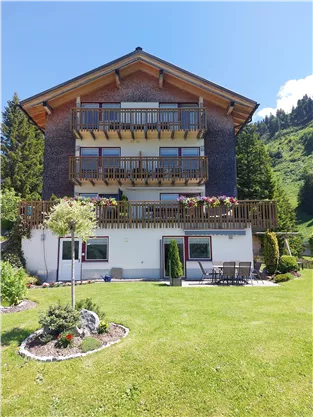 Ein schönes Haus mit mehreren Balkonen und einer gepflegten Gartenlandschaft. Die Umgebung ist von Bergen umgeben und strahlt Ruhe aus.