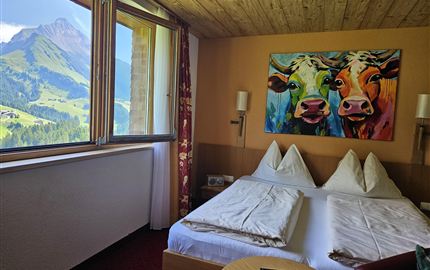 Een gezellig hotelkamer met twee bedden en een kleurrijke koe schilderij aan de muur. Door het raam is een indrukwekkend berglandschap te zien.
