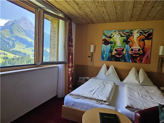 Ein gemütliches Hotelzimmer mit zwei Betten und einer farbenfrohen Kuhmalerei an der Wand. Durch das Fenster ist eine beeindruckende Berglandschaft zu sehen.