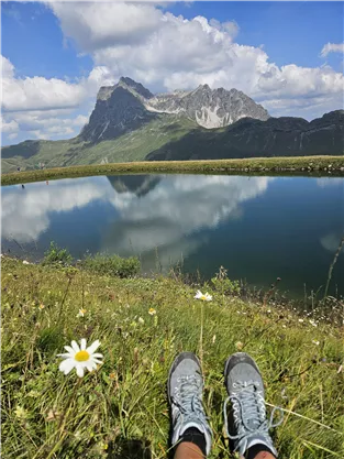 Ein schöner Bergsee mit reflektierenden Bergen im Hintergrund. Im Vordergrund sind Blumen und ein Paar Wanderschuhe zu sehen.