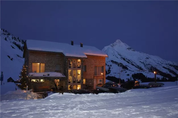 A cozy house in the mountains at night, surrounded by snow. The atmosphere is calm and inviting, with a sparkling light on the building.