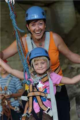 Eine Frau und ein Mädchen tragen Sicherheitsausrüstung und lächeln unter einem Felsen. Sie scheinen viel Spaß beim Klettern zu haben.