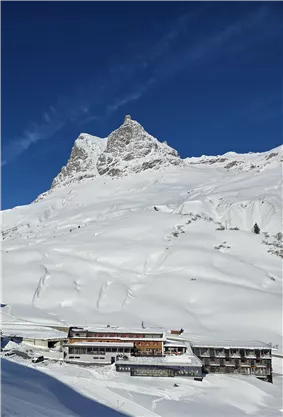 Ein schneebedecktes Gebirge mit einem beeindruckenden Gipfel im Hintergrund. Im Vordergrund steht ein gemütliches Gebäude, umgeben von einer winterlichen Landschaft.