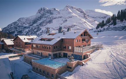 A picturesque mountain hotel in the snow with an outdoor pool. The surroundings are enveloped by snow-covered mountains, creating an idyllic winter landscape.