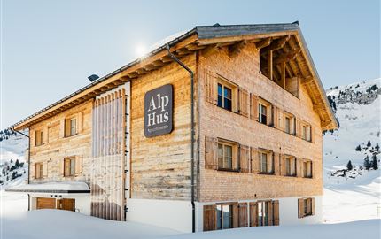 Ein gemütliches Holzhaus im alpinen Stil, umgeben von Schnee. Das Gebäude hat große Fenster und einen einladenden Eingang.