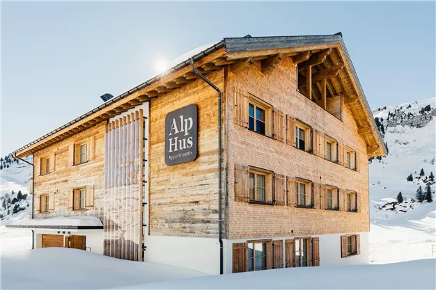 Ein gemütliches Holzhaus im alpinen Stil, umgeben von Schnee. Das Gebäude hat große Fenster und einen einladenden Eingang.