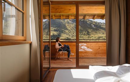 A cozy room with a large window. Outside, you can see an impressive mountain landscape and a person relaxing on the terrace.