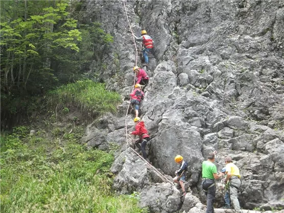 Eine Gruppe von Menschen klettert an einem Felsen mit Seilsicherung. Im Hintergrund sind grüne Pflanzen und Bäume zu sehen.