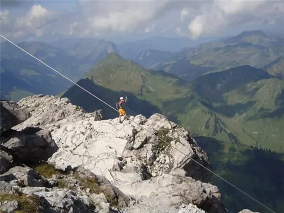 A climber stands on a rocky mountain peak and enjoys the view. In the background, green, forested mountains and a clear sky can be seen.