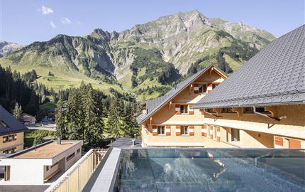Eine malerische Berglandschaft mit hohen Bergen und grünen Wäldern. Im Vordergrund ist ein Schwimmbad, das sich harmonisch in die Umgebung einfügt.