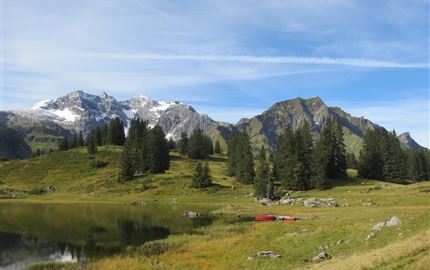 Eine malerische Berglandschaft mit schneebedeckten Gipfeln und grünem Gras. Im Vordergrund ist ein ruhiger See zu sehen.