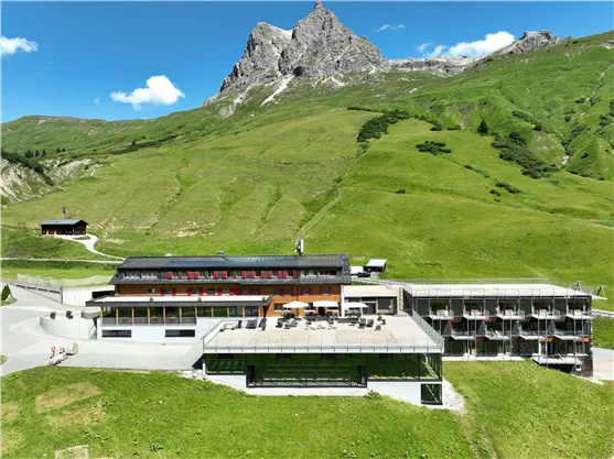 Ein modernes Hotel liegt inmitten einer grünen Berglandschaft. Im Hintergrund sind majestätische Berge und ein klarer blauer Himmel zu sehen.