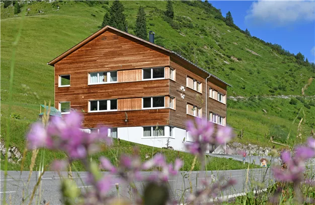 A wooden house surrounded by green hills and colorful flowers. The sky is clear and blue.