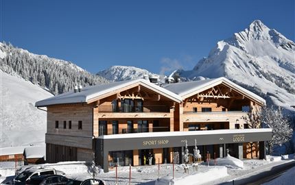 Ein modernes Gebäude in den Bergen, umgeben von Schnee. Im Vordergrund sind Parkplätze und eine Sport-Shop-Anzeige sichtbar.