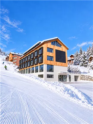 A modern hotel in a snow-covered landscape. The sky is blue and the slope is well-groomed.
