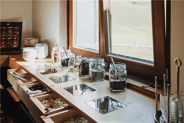 Ein Frühstückstisch mit Gläsern voller Müsli und Snacks. Große Fenster lassen viel Licht herein und bieten einen Blick nach draußen.