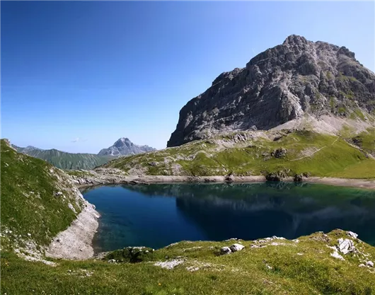 Ein klarer Bergsee umgeben von grünen Wiesen und majestätischen Bergen. Der Himmel ist blau und wolkenlos.