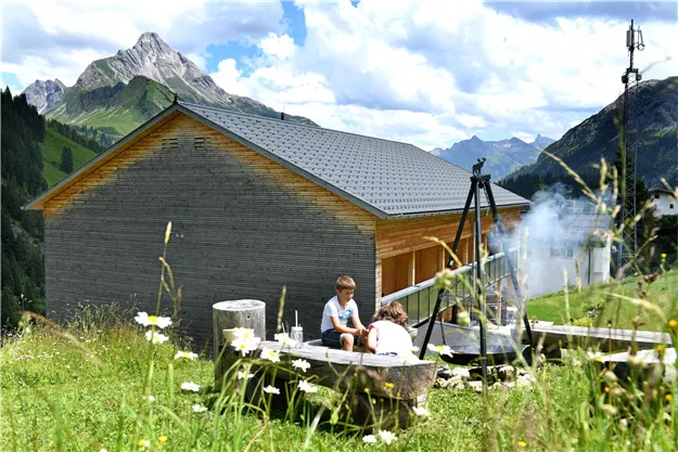A cozy mountain cabin in nature, surrounded by green meadows and mountains. Two children are playing near a barbecue area.