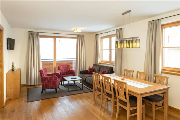 Ein gemütliches Wohnzimmer mit einem Esstisch und modernen Möbeln. Helle Fenster lassen viel Tageslicht herein und schaffen eine freundliche Atmosphäre.