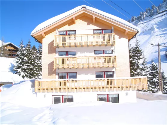 A cozy wooden building with several balconies in a snow-covered landscape. The sun is shining, and there are low mountains visible in the background.