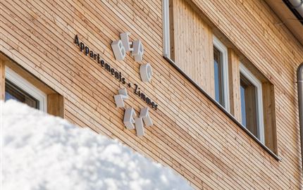 Ein Holzhaus mit einem Schild, das „Apartments & Zimmer“ zeigt. Der Schnee liegt im Vordergrund und verleiht der Szene eine winterliche Atmosphäre.