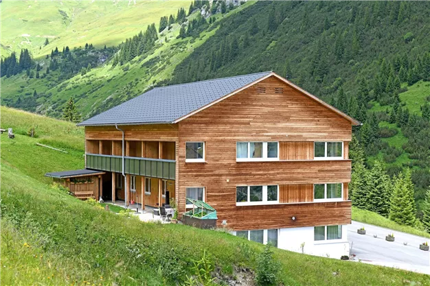 A cozy wooden house in the midst of a green landscape. The surroundings are hilly and covered with trees.