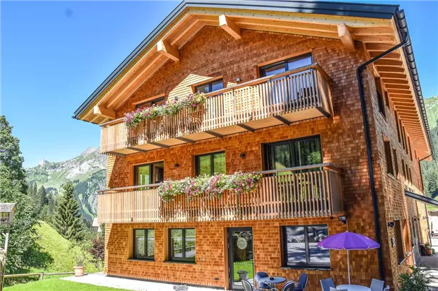 Ein gemütliches Holzhaus mit mehreren Balkonen und Blumenkästen. Im Hintergrund sind die Berge und ein klarer blauer Himmel zu sehen.